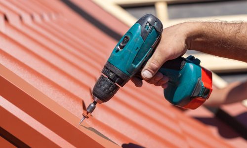 Worker on a roof with electric drill installing red metal tile on wooden house
